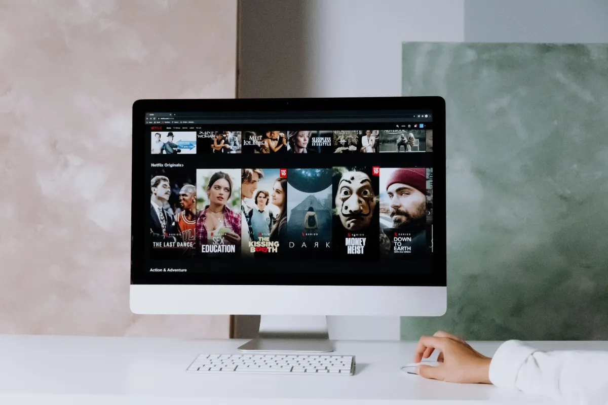 A desktop computer on a white desk showing rows of film and series artwork in a streaming catalogue, with a hand resting on the mouse