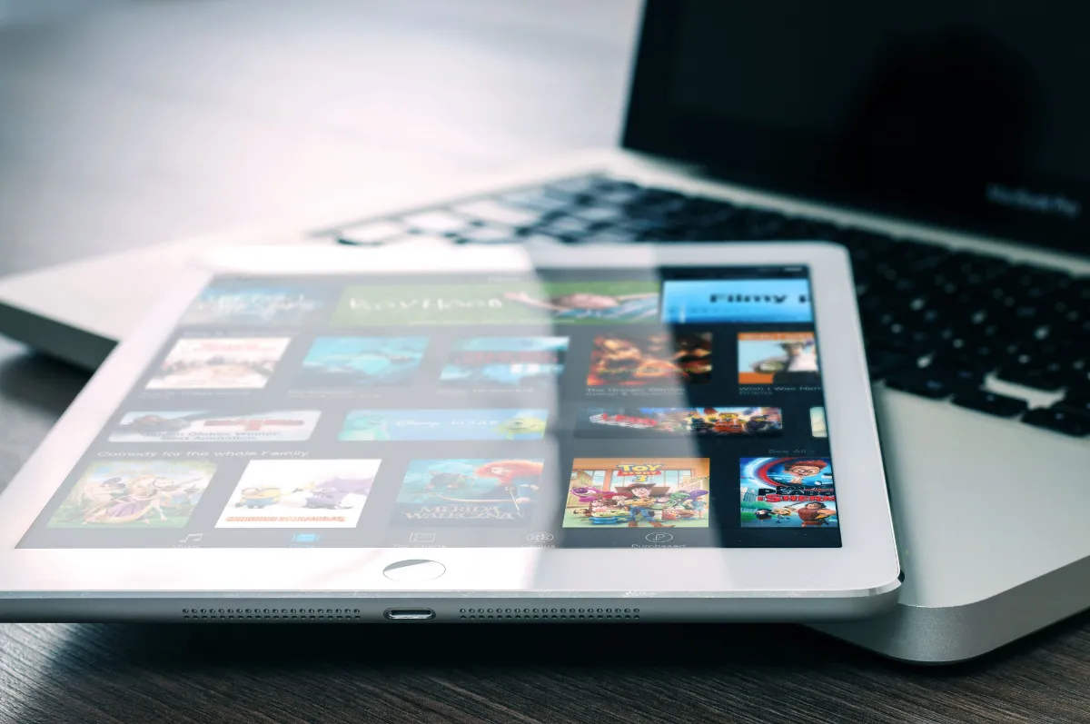 A tablet lying on the keyboard of an open laptop, its screen filled with a grid of film and television thumbnails from a video library